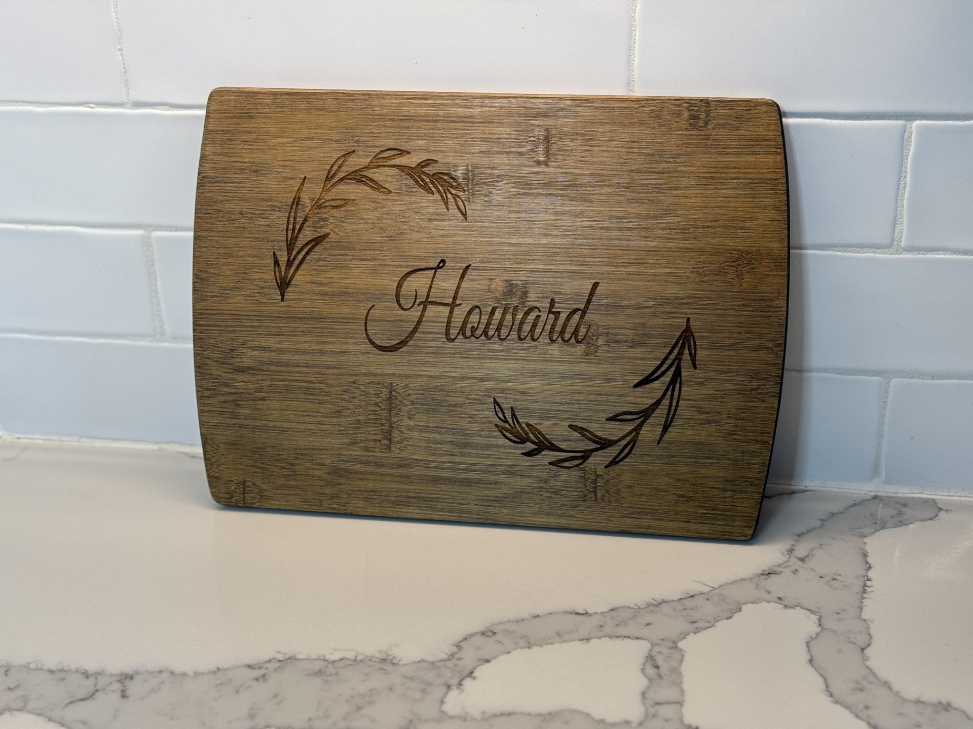 The Personalized Family Information Cutting Board showcases the name Howard elegantly engraved in the center, with decorative leaf motifs at the top left and bottom right corners, resting on a white marble countertop with gray veining that complements the tiled backdrop.