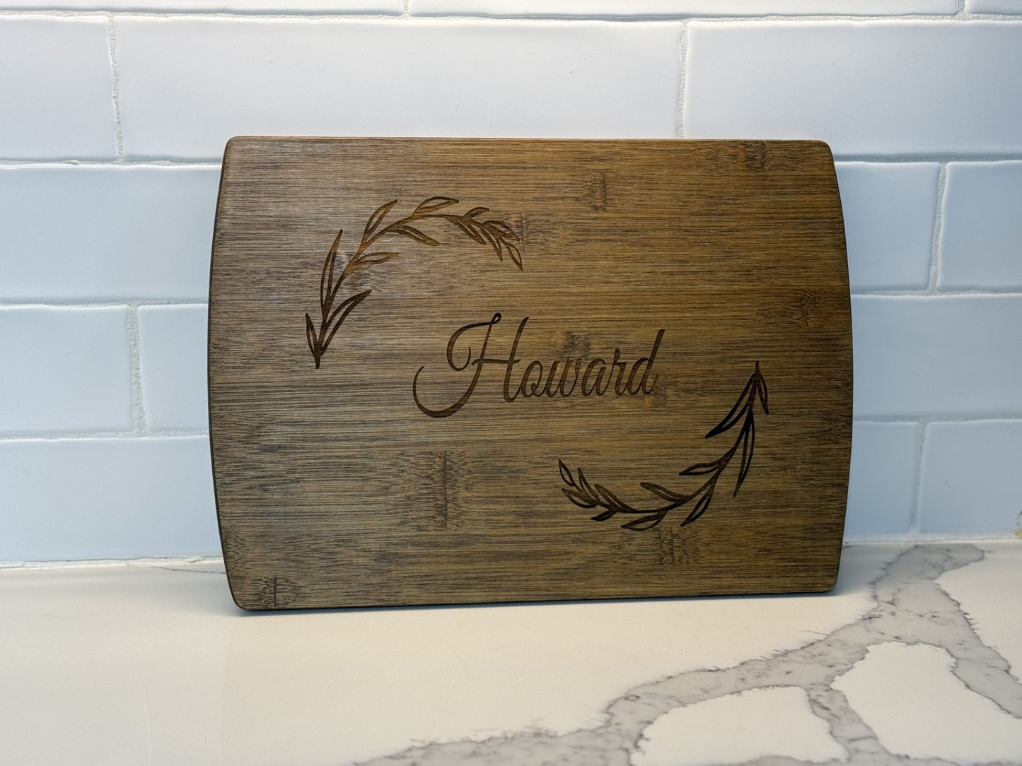 A Personalized Family Information Cutting Board displays Howard elegantly engraved at the center with leafy branches on either side, resting against a white tile backsplash and gray-veined marble surface.