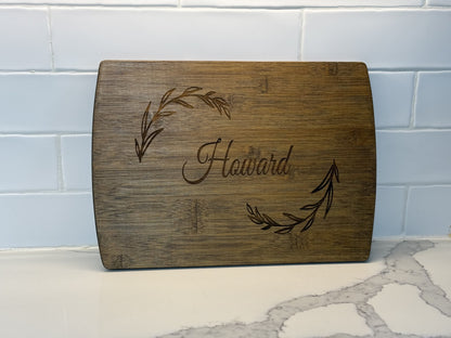 A Personalized Family Information Cutting Board displays Howard elegantly engraved at the center with leafy branches on either side, resting against a white tile backsplash and gray-veined marble surface.