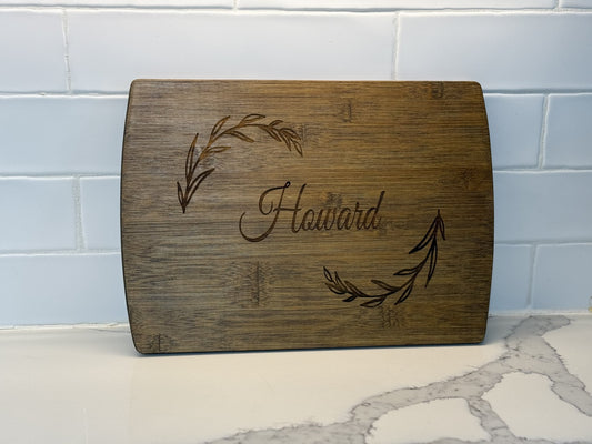 A personalized family information cutting board with Howard engraved in the center, flanked by two leaf designs, rests on a white marble counter against a white tiled backsplash.