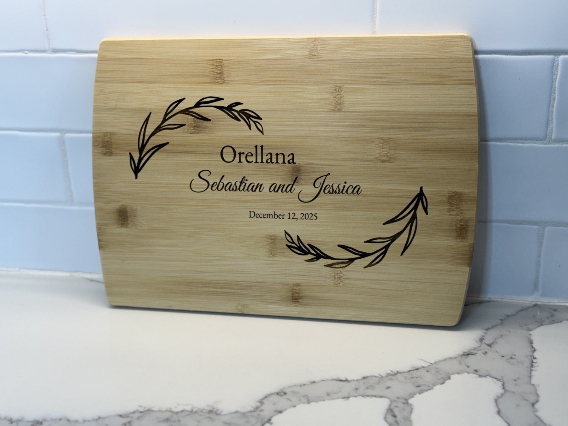 The Personalized Family Information Cutting Board, engraved with Orellana, December 12, 2025, features decorative laurel branches. It rests elegantly against a white tiled wall on a sleek marble countertop.