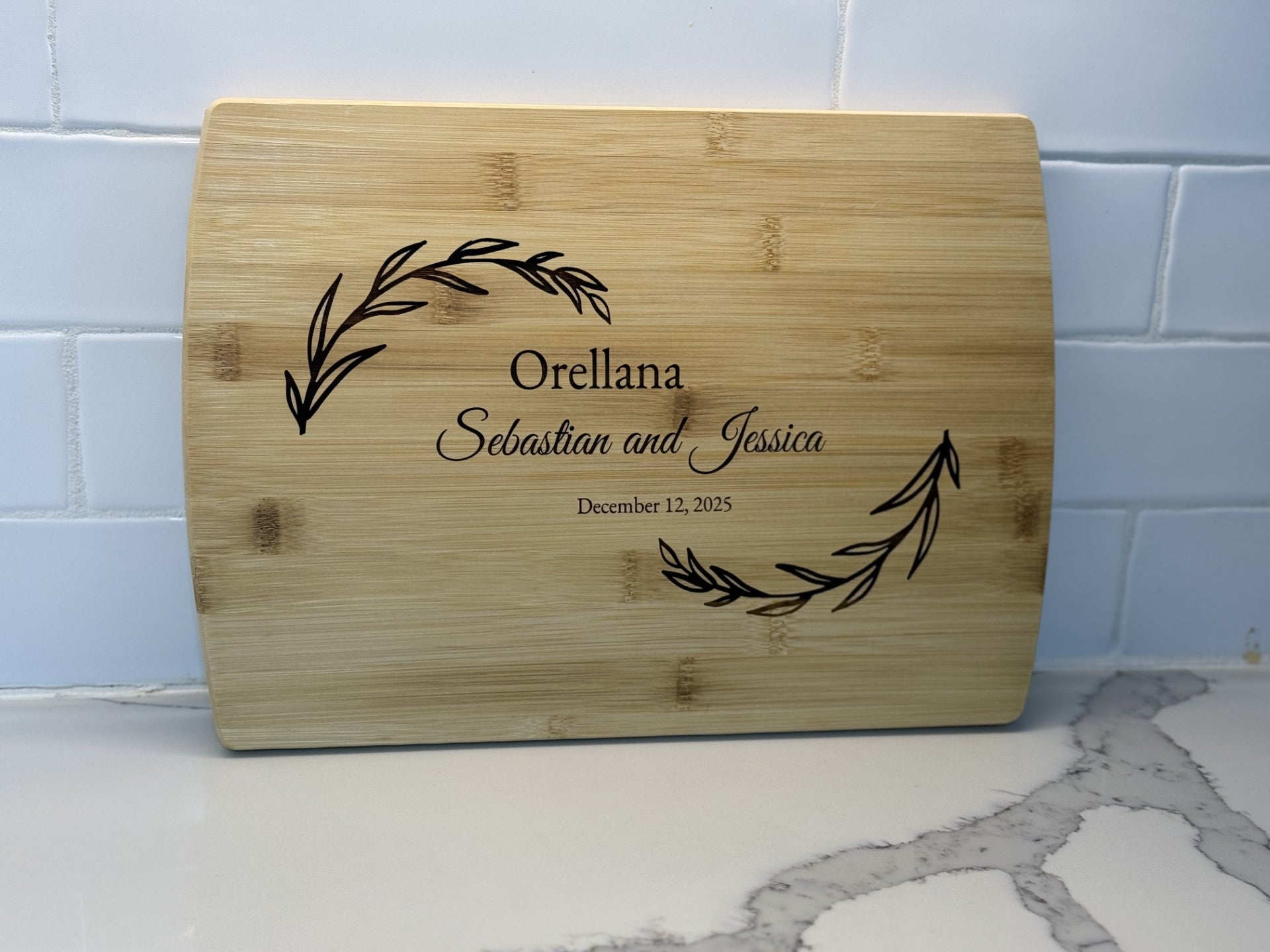 A personalized wooden cutting board is engraved with Orellana, and December 12, 2025, surrounded by decorative leaf designs. Its propped against a white tiled wall on a gray-veined countertop. Product: Personalized family information cutting board.