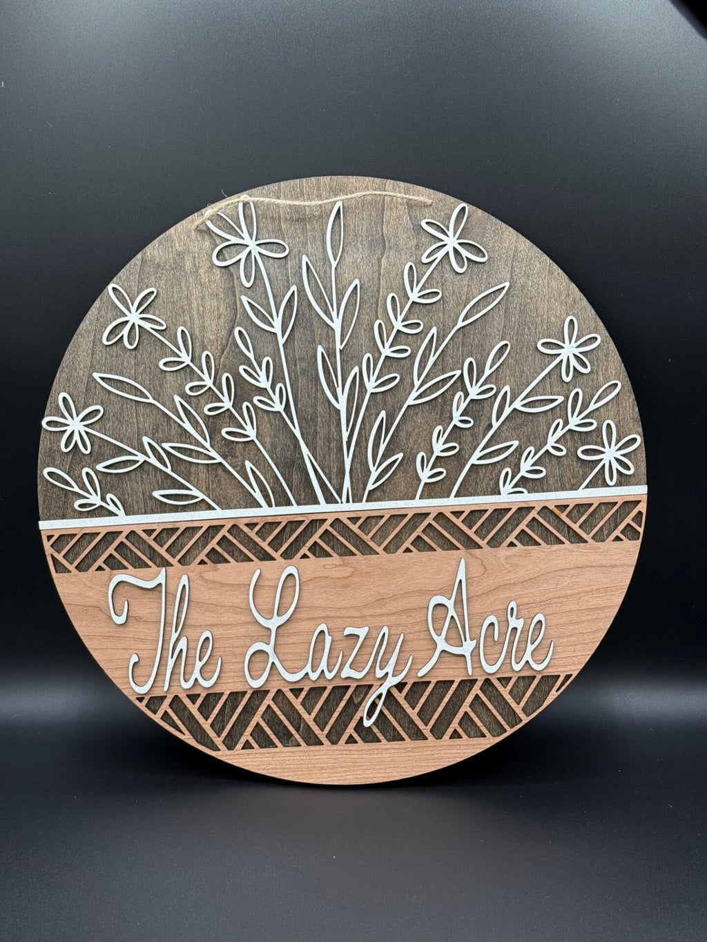 The Personalized Floral Basket Welcome Sign Door Hanger features elegant cursive The Lazy Acre on dark woodgrain, with decorative white floral outlines. Its oval shape and diamond pattern exude warmth and style, offering customizable and charming décor.