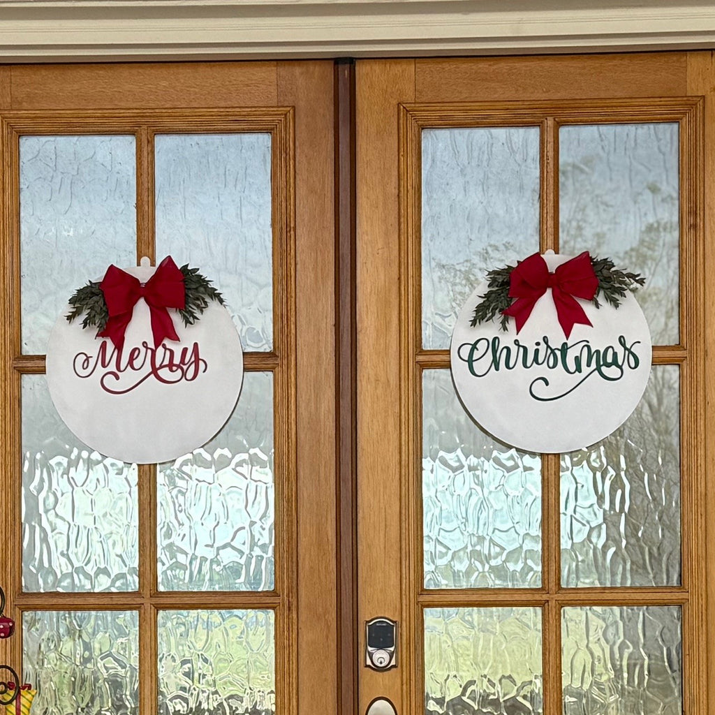 A set of wooden double doors decorated for Christmas. Each door has a round white wreath with a red bow; the left wreath says Merry, and the right one says Christmas. A plant and decorative stand sit to the right, with a reindeer statue to the left, in front of textured glass panels.
