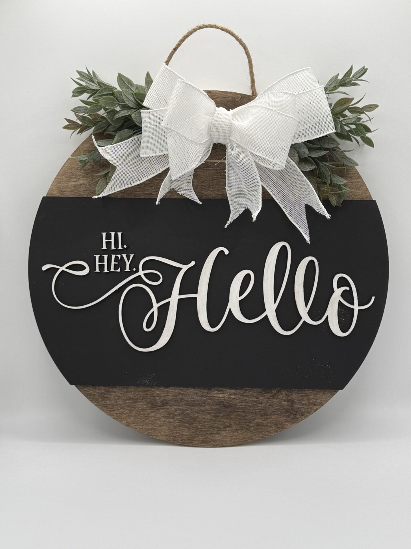 Round wooden sign with a black center featuring the words Hi. Hey. Hello in white cursive. Decorated with a large white bow at the top and surrounded by green foliage. A natural string loop is attached for hanging.