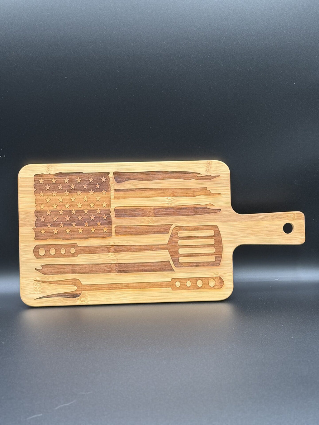 A wooden cutting board with a handle features etched designs of barbecue utensils and the American flag. The utensils include knives, forks, and a spatula, creatively aligned to form the flags stripes and field of stars. The board is set against a plain dark background.