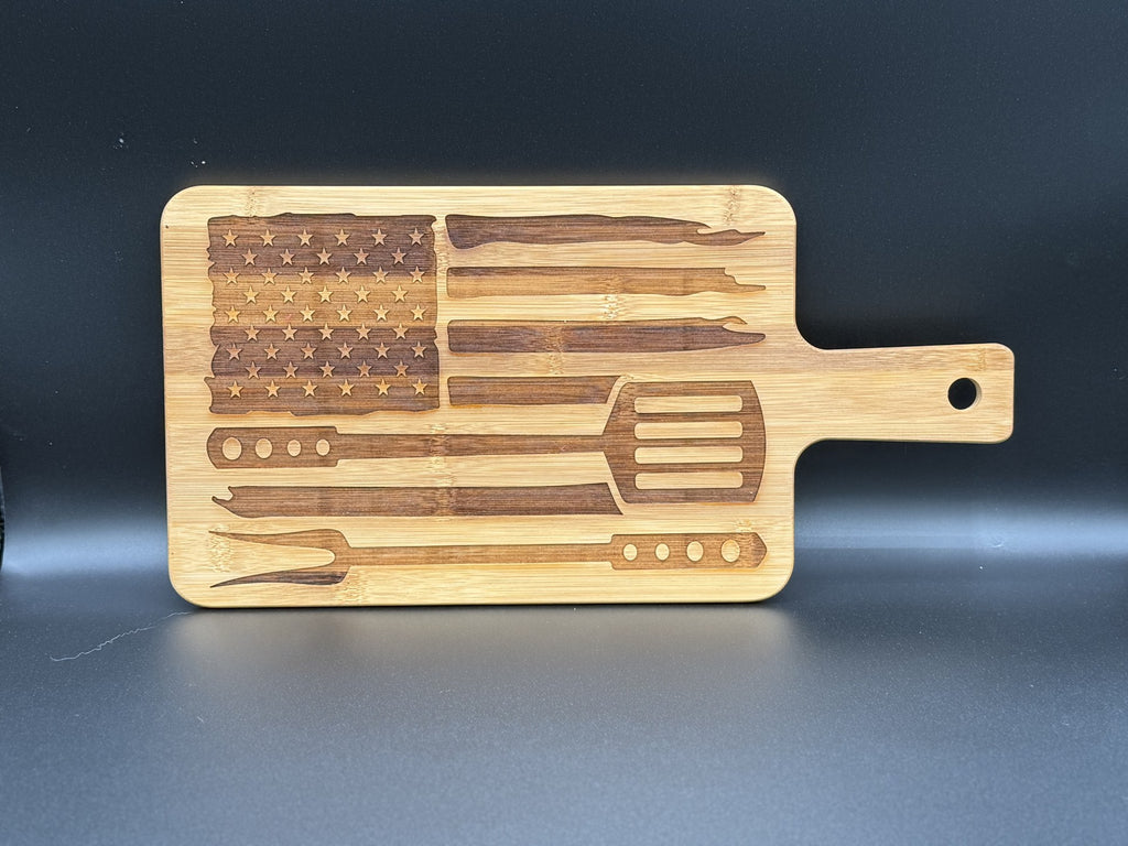 A wooden cutting board features an engraved design of the American flag using grilling tools. The flags field of stars is made of a fork pattern, and the stripes are represented by various grilling utensils, including spatulas and skewers. The board includes a handle with a hole.