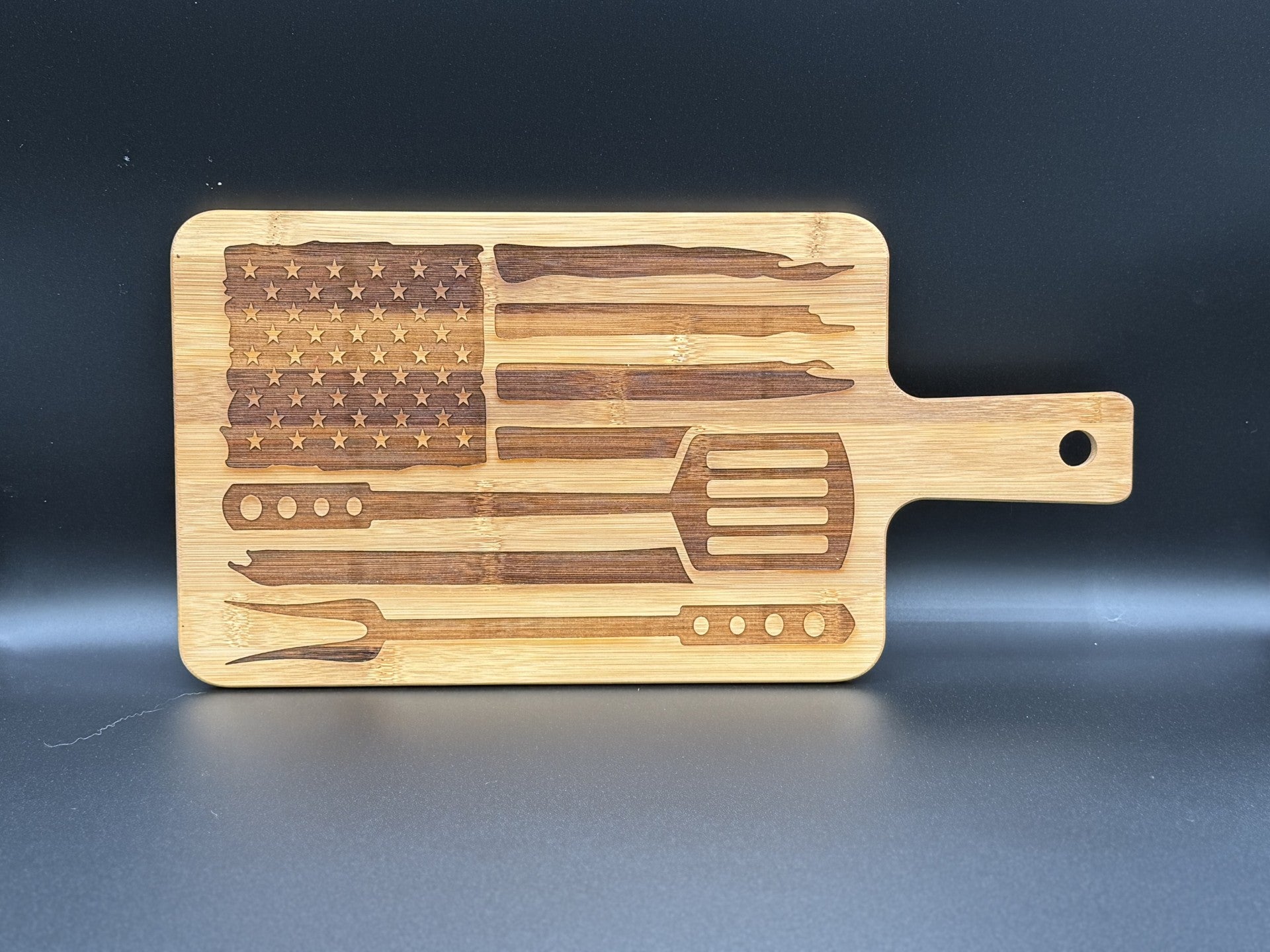 A wooden cutting board with an engraved design featuring an American flag pattern. The flag is made from various grilling tools, including a spatula, fork, tongs, basting brush, and skewer. The board has a handle on the right side and is set against a dark background.