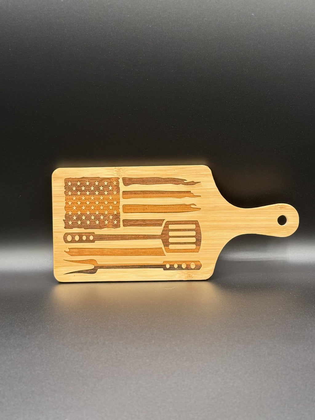A wooden cutting board features a design resembling the American flag, with the flags stripes formed by grilling utensils like a spatula, tongs, and skewers. The stars are on the left side, with a perforated handle on the right. The board is set against a dark background.