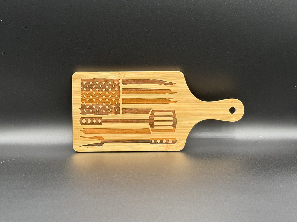 Wooden cutting board with an integrated handle, featuring an engraved design. The engraving includes the American flag and various barbecue utensils such as a fork, spatula, and tongs arranged horizontally. The board is set against a plain dark background.