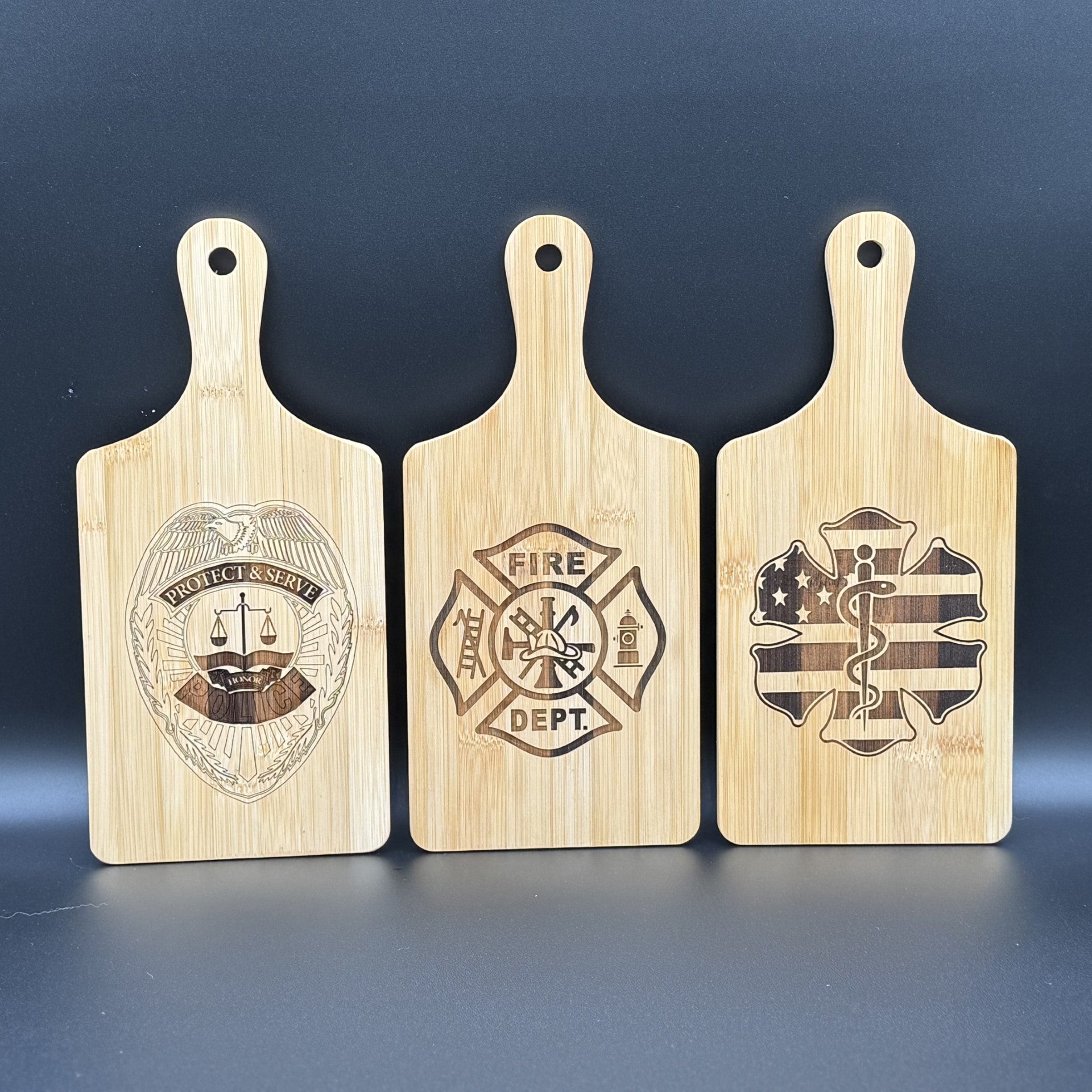 Three Military Logo Cutting Boards with handles are displayed vertically against a dark background. Each cutting board features engravings of a police badge, fire department emblem, and star of life symbol from left to right, honoring those who protect and serve.