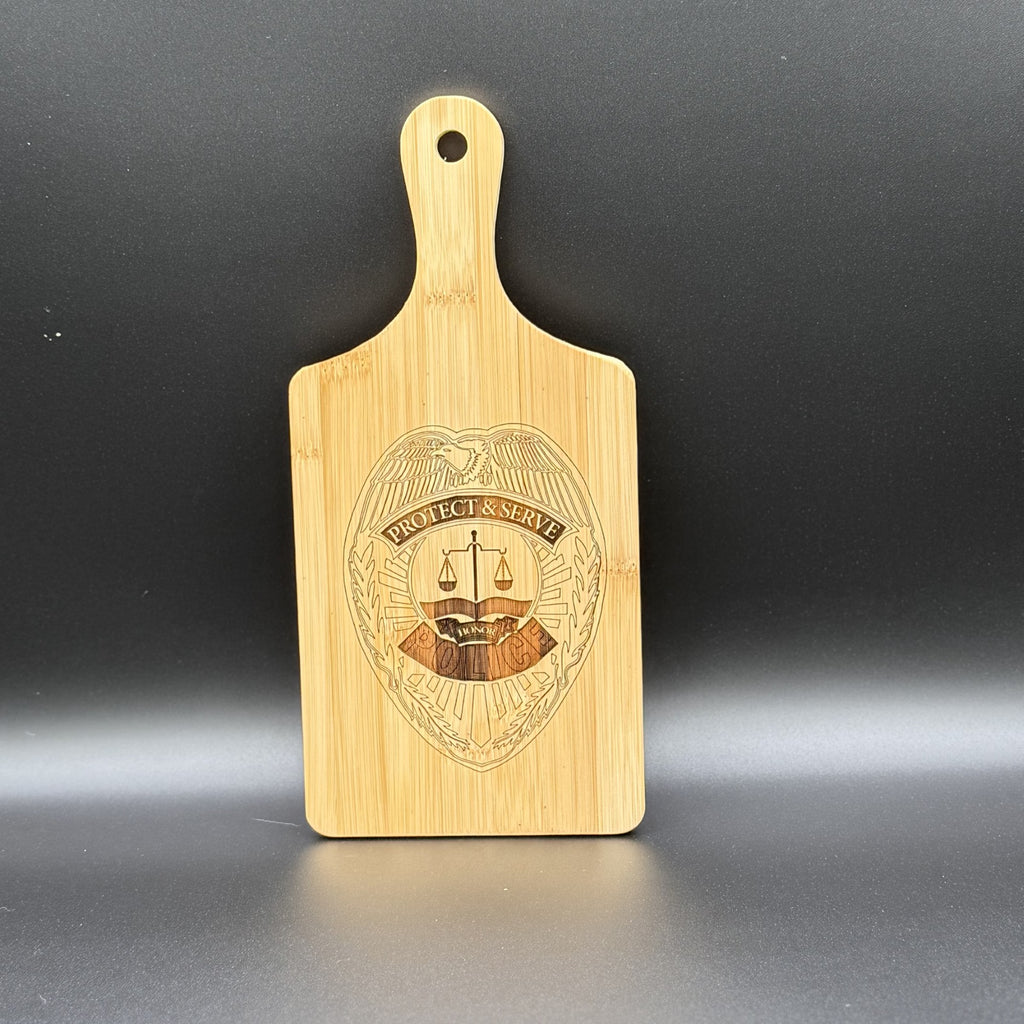 The Military Logo Cutting Board features a handle and an engraved shield with an eagle, PROTECT & SERVE, and scales of justice. The light wood against a dark backdrop beautifully highlights its intricate texture and design.