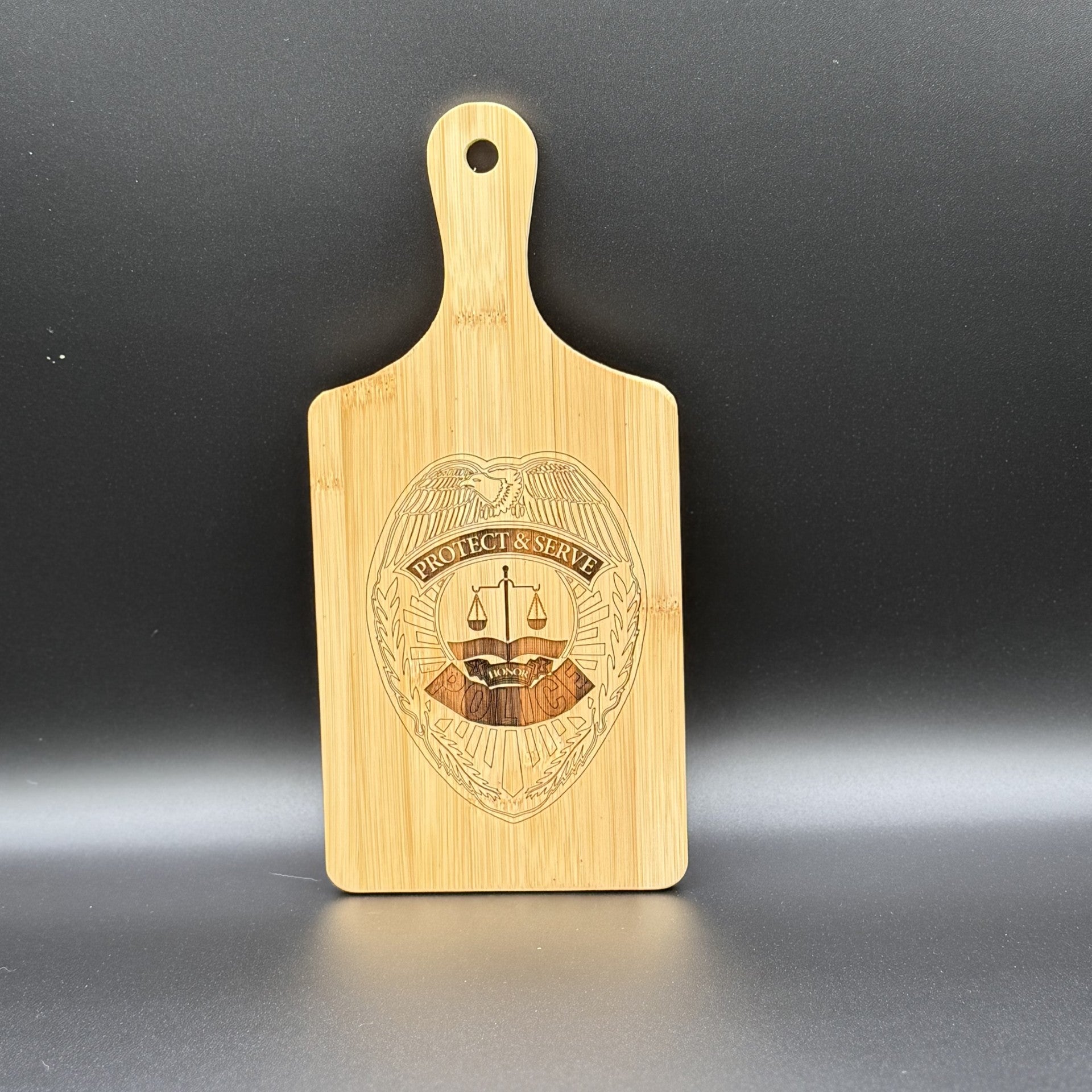 The Military Logo Cutting Board features a handle and an engraved shield with an eagle, PROTECT & SERVE, and scales of justice. The light wood against a dark backdrop beautifully highlights its intricate texture and design.