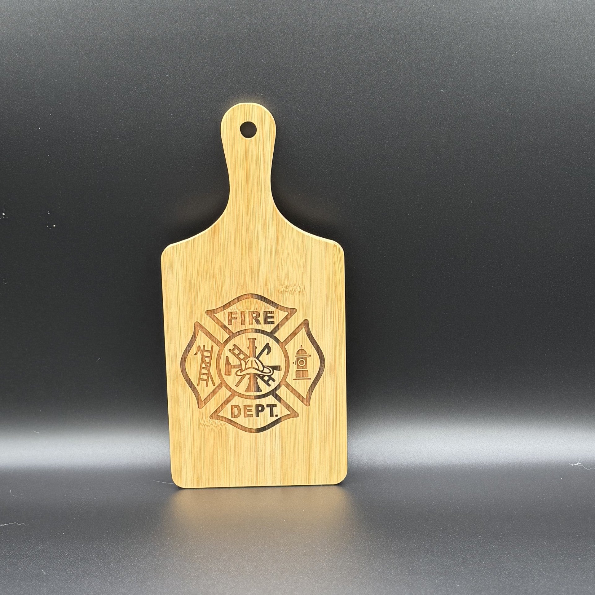 A wooden military logo cutting board with a handle features an engraved firefighter emblem and logo, including a ladder, helmet, fire hydrant, and axe around FIRE DEPT., elegantly displayed against a dark gray backdrop.