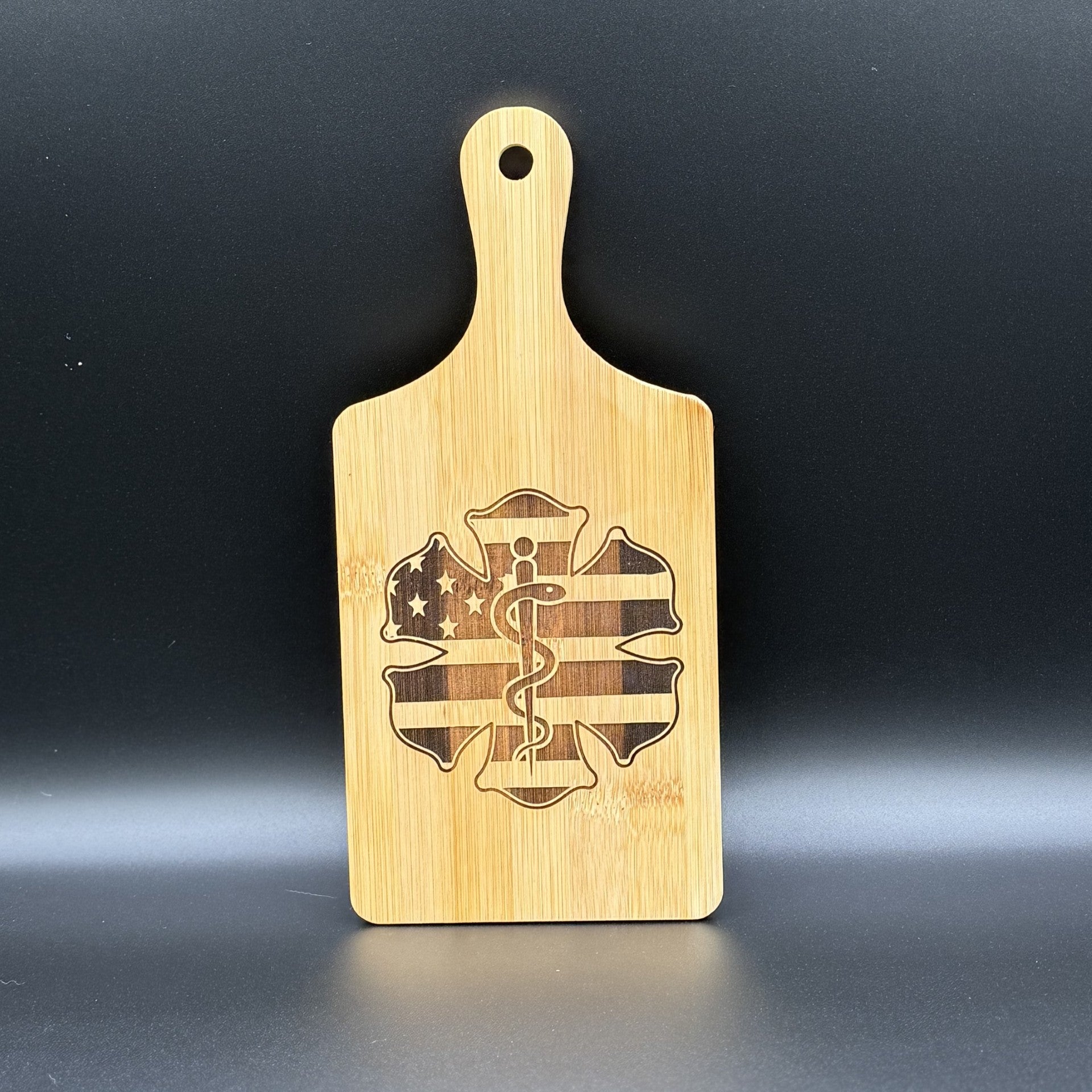 The Military Logo Cutting Board is a bamboo board with a handle, featuring a star of life logo with an American flag design combining stars and stripes, set against a plain black background. Perfect for military enthusiasts.