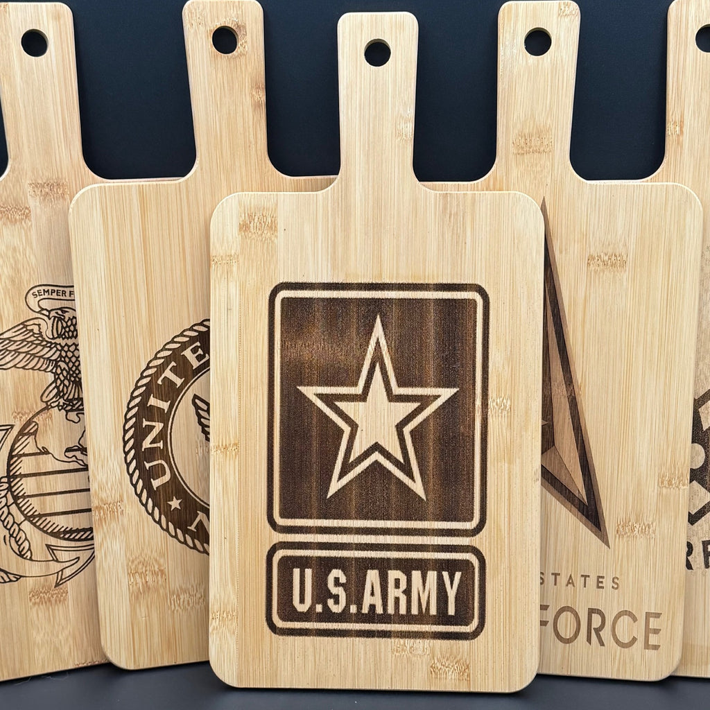 Five wooden cutting boards with handles are displayed upright. Each board features a different engraved U.S. military emblem. The central board prominently displays the U.S. Army emblem with a star and text. Surrounding boards show partial emblems of the other military branches.