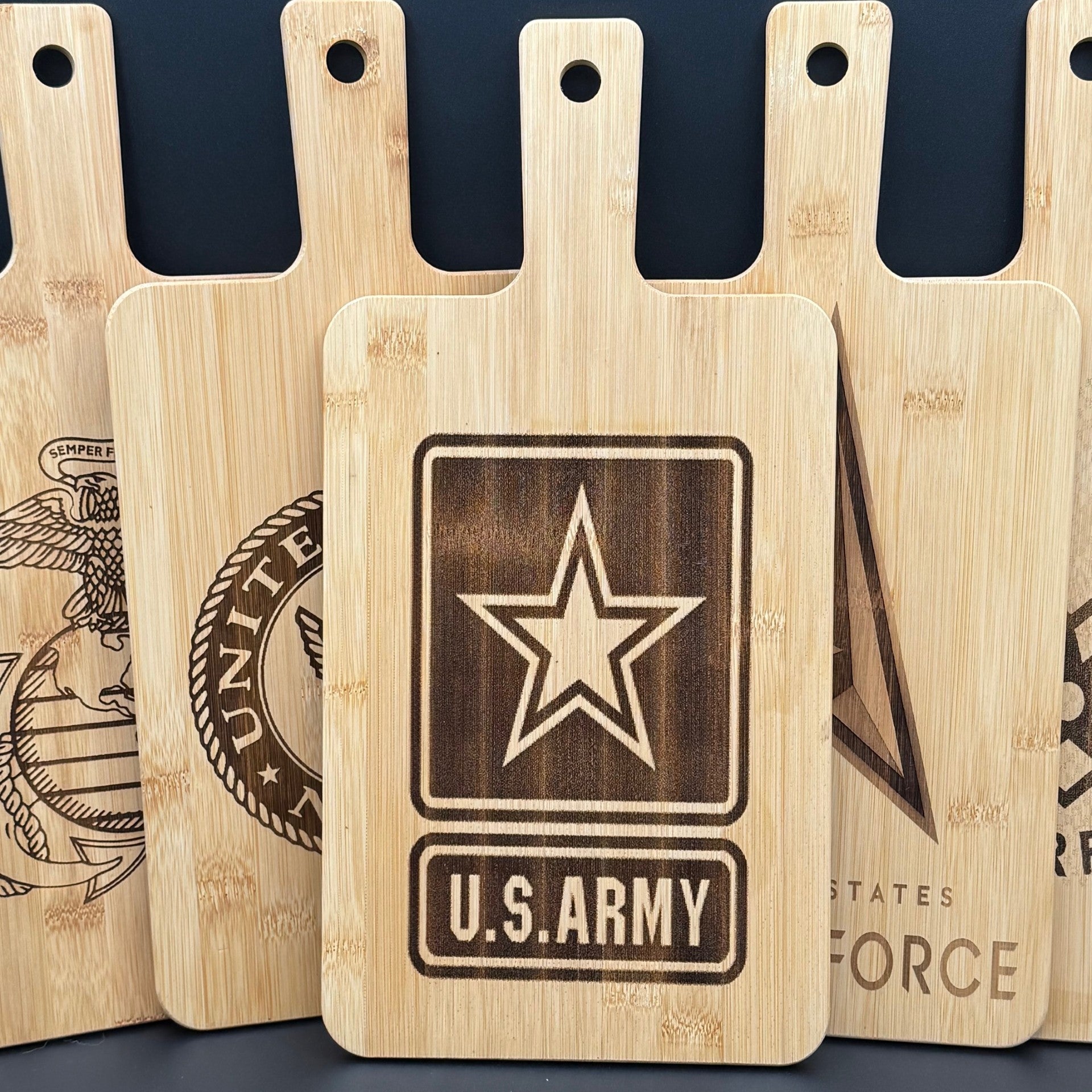 Five wooden cutting boards with handles are displayed upright. Each board features a different engraved U.S. military emblem. The central board prominently displays the U.S. Army emblem with a star and text. Surrounding boards show partial emblems of the other military branches.