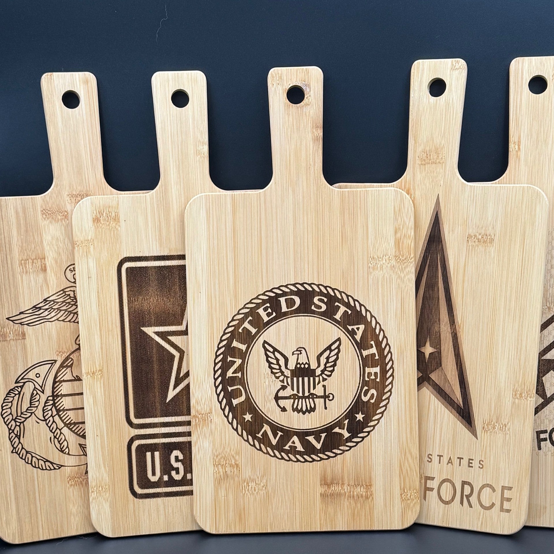Five wooden cutting boards are displayed upright. Each board features a different carved emblem: U.S. Navy, a star, and others partially visible. They have handles with holes for hanging. The boards are made of light-colored wood, showcasing natural grain patterns.