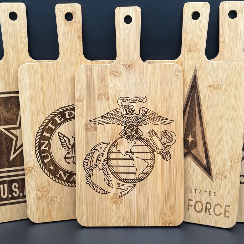 Five wooden cutting boards are arranged vertically. The central board features a detailed engraving of an eagle perched on a globe with an anchor, and a banner reading Semper Fidelis. Surrounding boards display different U.S. military insignias, including the Navy and Air Force.