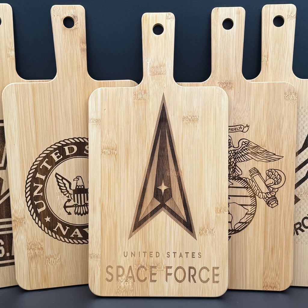 Five wooden cutting boards are displayed upright. Each board has a different emblem engraved on it, including the United States Space Force logo prominently shown in the center. The boards have a light wood color and elongated handles with hanging holes.