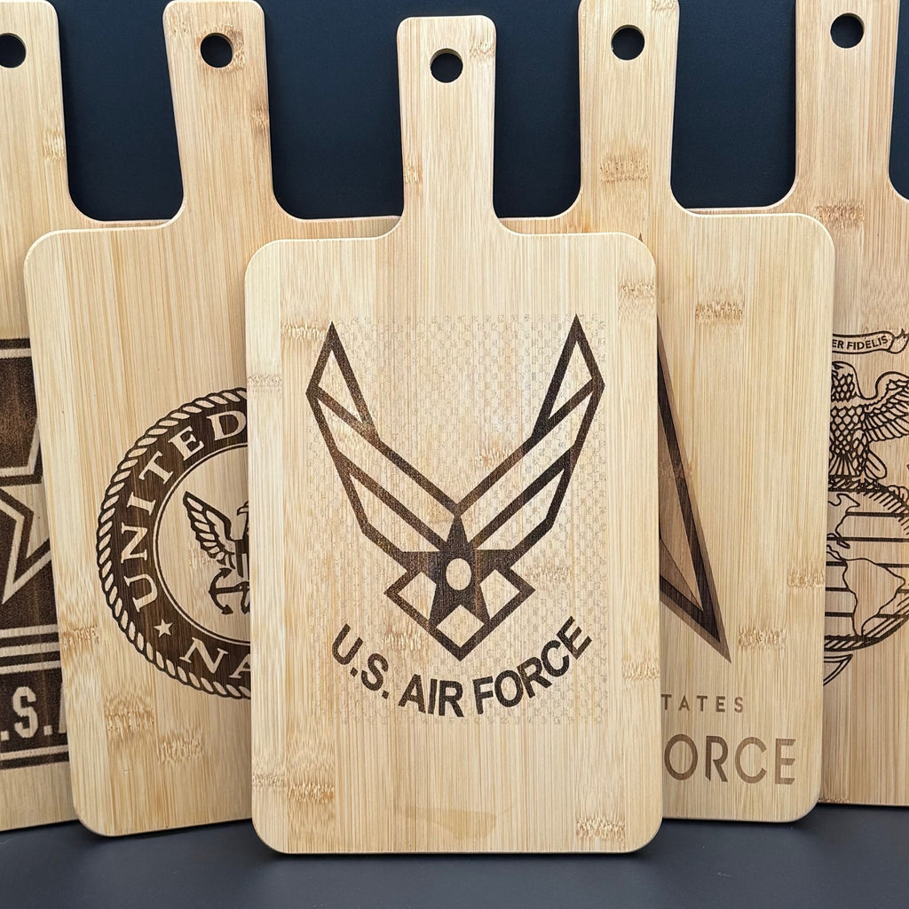 A collection of wooden cutting boards displays various etched military logos. The central board prominently features the U.S. Air Force logo and text. The boards are arranged upright with handles pointing upwards, showcasing their detailed engravings against a dark background.