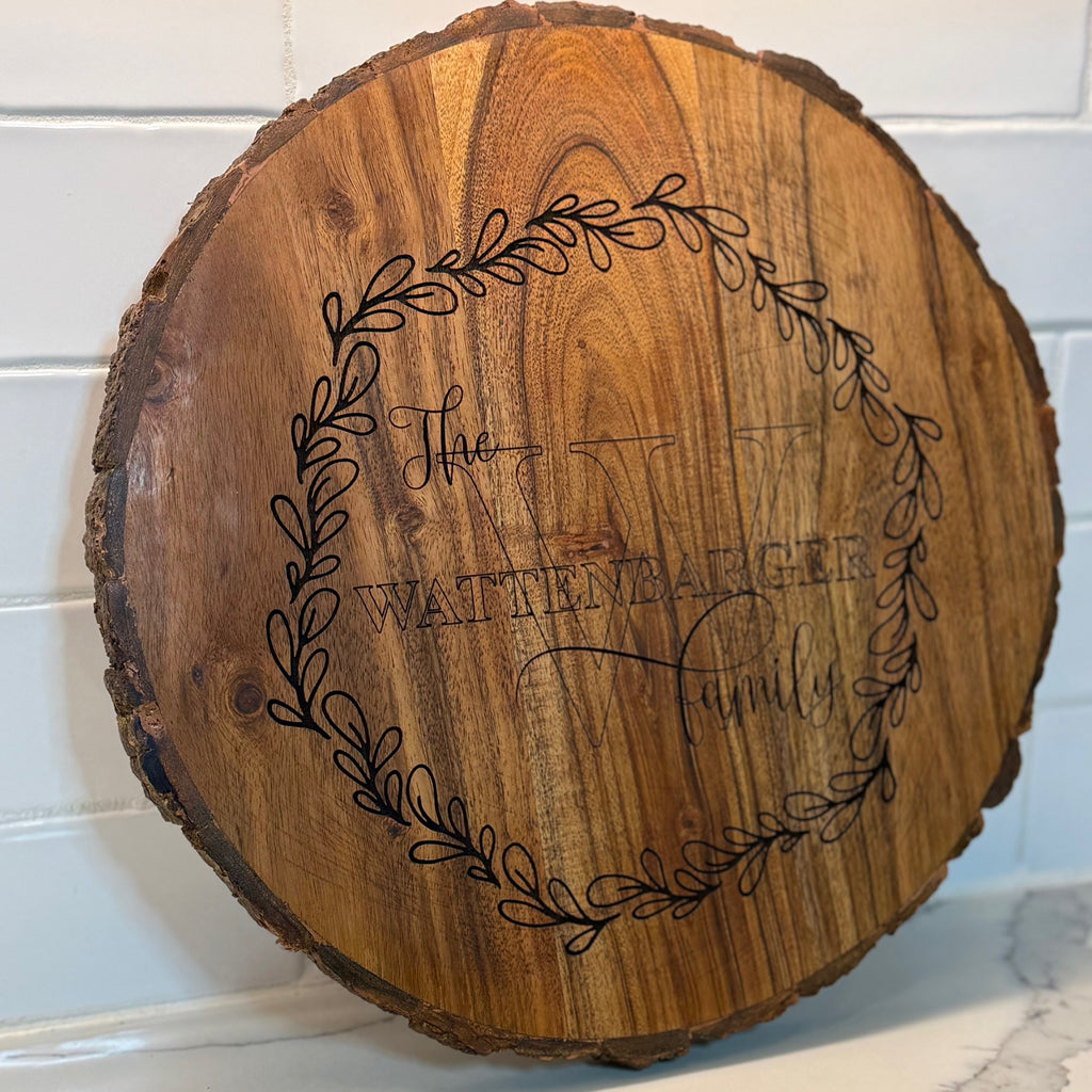 A round wooden plaque with a natural bark edge, featuring an engraved decorative wreath surrounding the text The Wattenbarger Family prominently displayed in the center. It rests against a white tiled background.