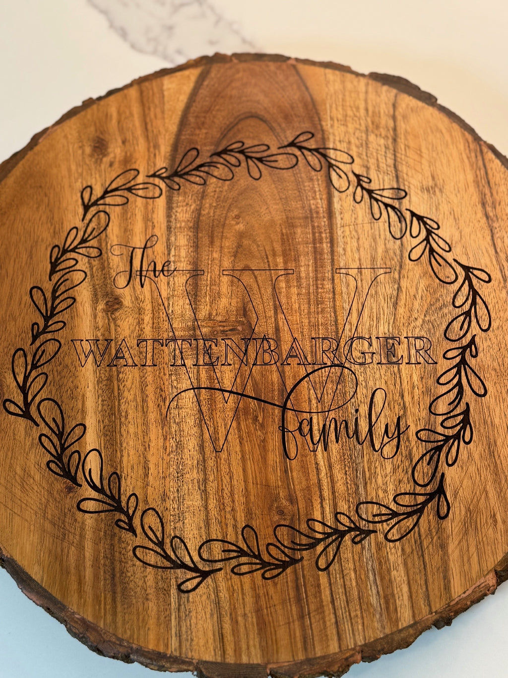 A round wood slice features the text The Wattenbarger Family engraved in the center with a large decorative W behind it. A leafy wreath design encircles the text on the woods surface, highlighting the natural grain.