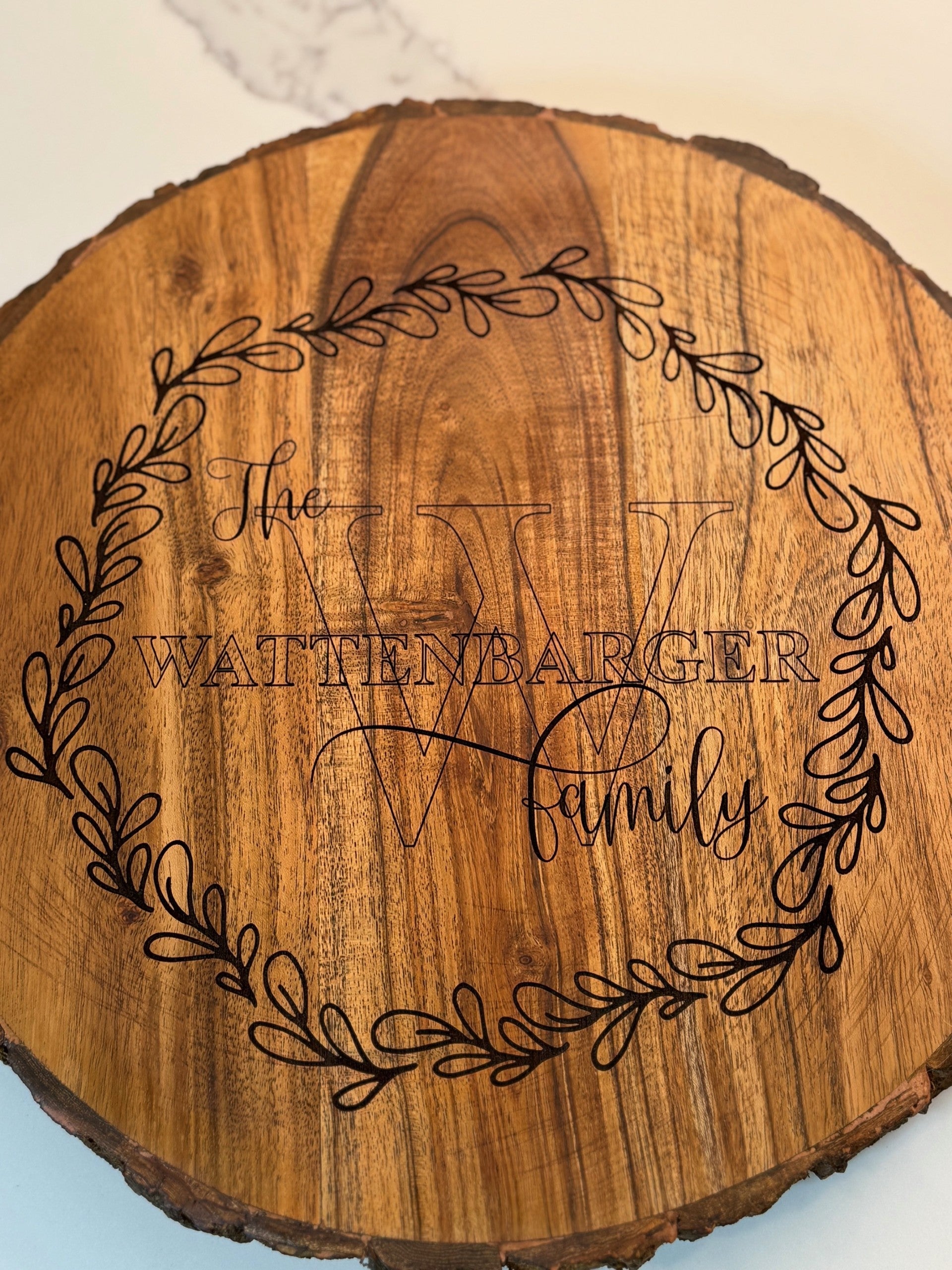 A round wood slice features the text The Wattenbarger Family engraved in the center with a large decorative W behind it. A leafy wreath design encircles the text on the woods surface, highlighting the natural grain.