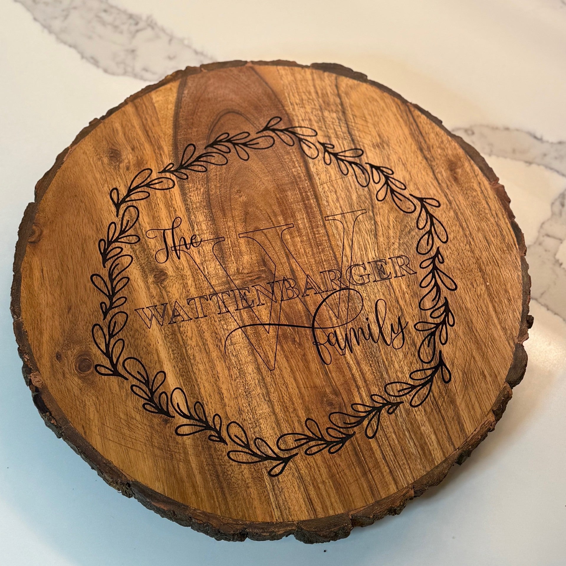 A round wooden plaque with bark edges sits on a marble surface. It features an engraved wreath design encircling the words The Wattenbarger Family with a large letter W in the background. The wood has a rich, natural grain pattern.