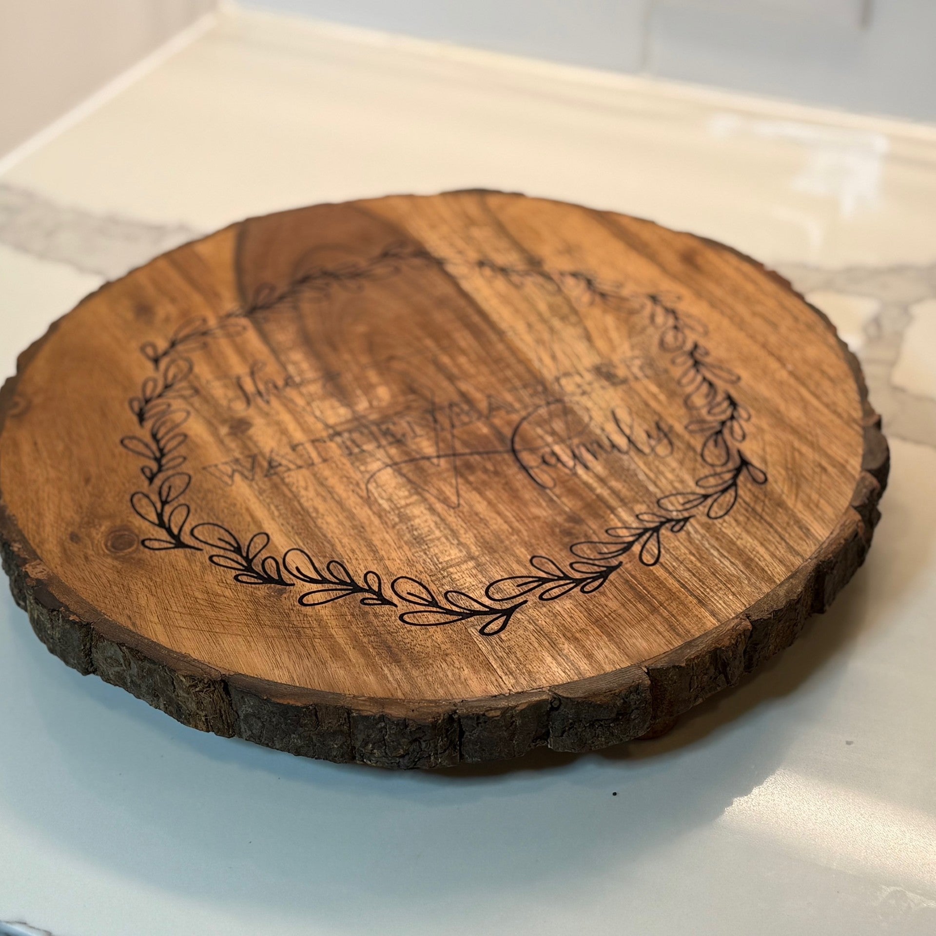 A round wooden serving board with bark edges sits on a counter. The board features an engraved design with a laurel wreath encircling the text: The Walters Family. The wood has a smooth finish, and the counter has a marble-like appearance.