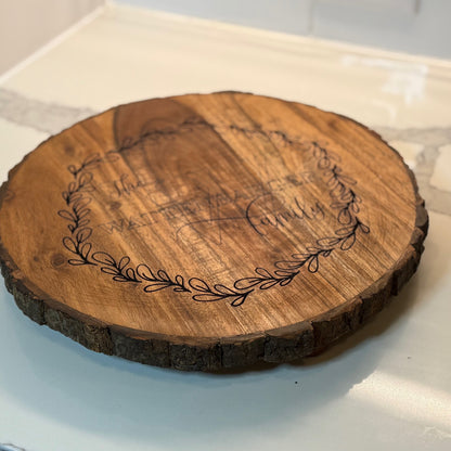 A round wooden serving board with bark edges sits on a counter. The board features an engraved design with a laurel wreath encircling the text: The Walters Family. The wood has a smooth finish, and the counter has a marble-like appearance.