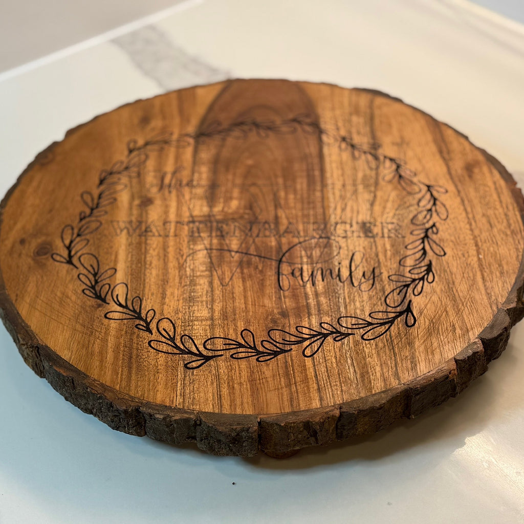 A round wooden board with bark edges features a laser-engraved design. In the center, it says The [NAME] Family surrounded by a delicate leaf wreath. The wood has a rich grain and the board sits on a smooth white surface.