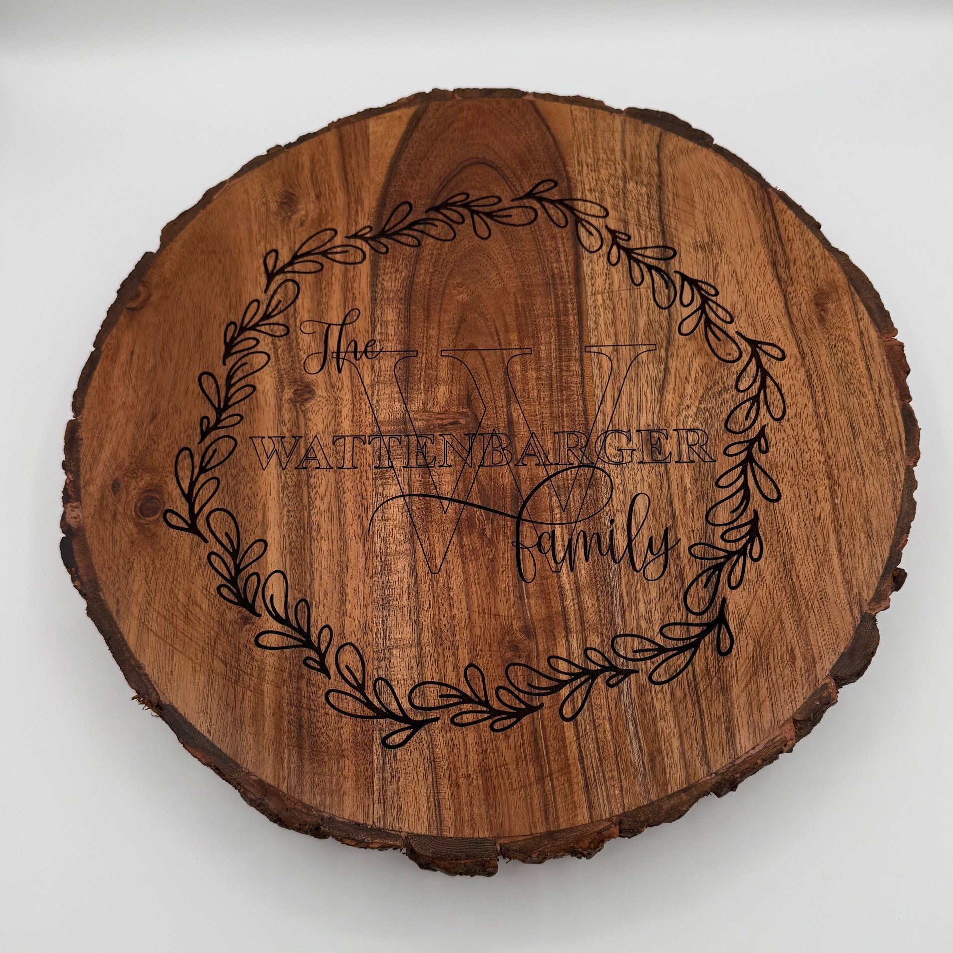 A round wooden plaque with bark edges features an engraved design. In the center, a large letter W is overlaid by text reading The Wattenbarger Family. A decorative wreath of leaves encircles the text, forming its border. The wood has a natural, polished finish.