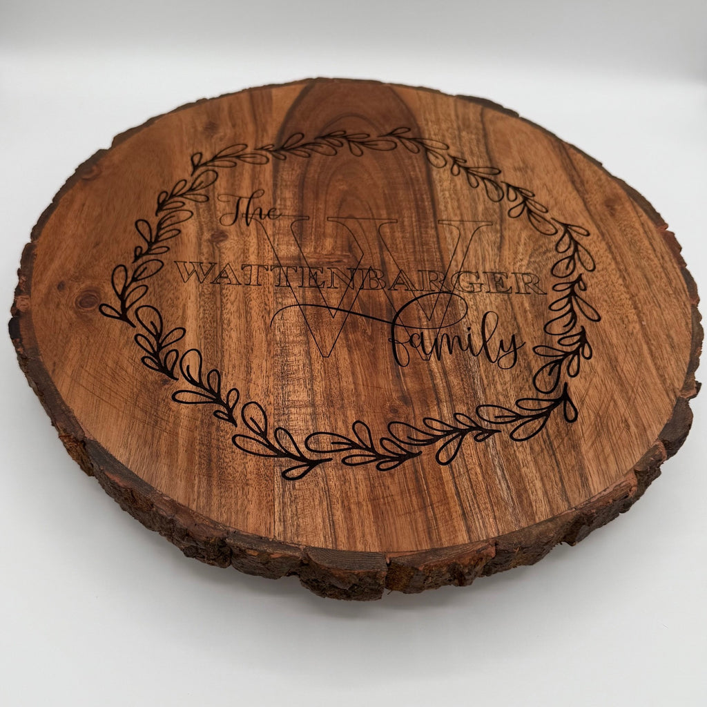 A round wooden plaque with bark edging features an engraved wreath design. Inside the wreath are the words The Wattenbarger Family with an elegant script font for Family. The plaque has a rustic appearance with the grain of the wood visible.