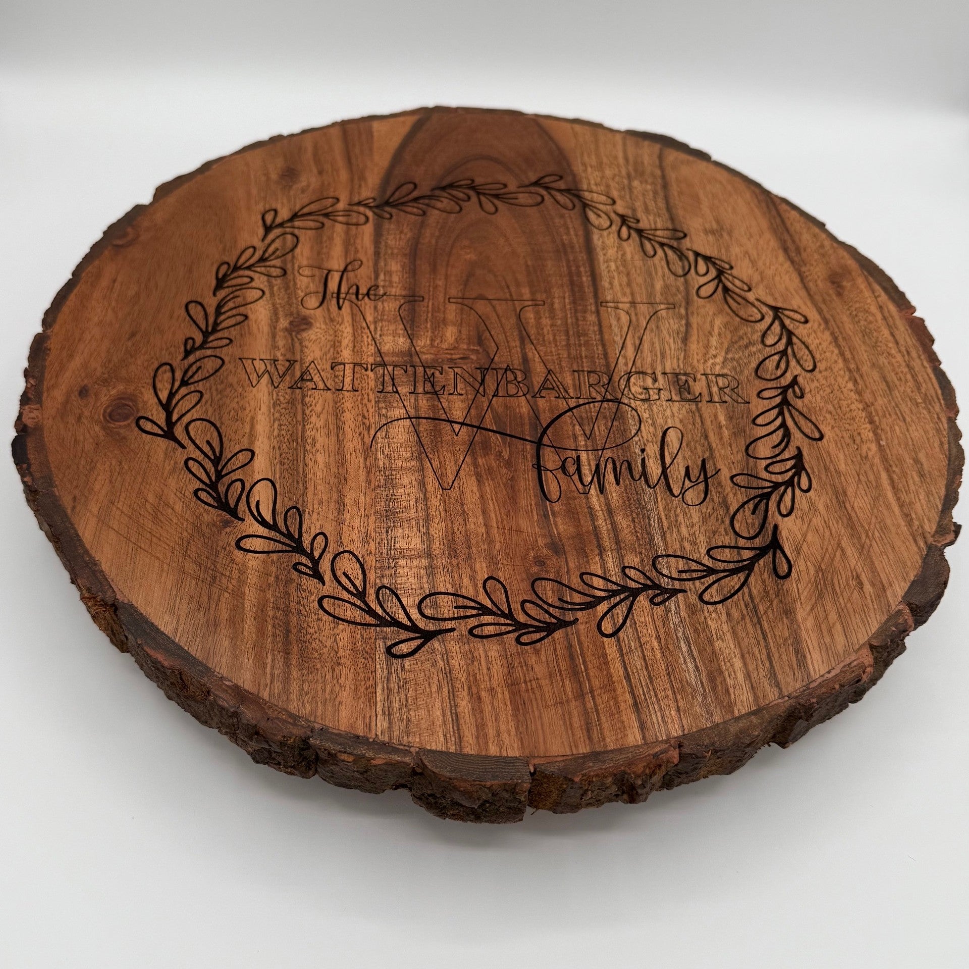 A round wooden plaque with bark edging features an engraved wreath design. Inside the wreath are the words The Wattenbarger Family with an elegant script font for Family. The plaque has a rustic appearance with the grain of the wood visible.
