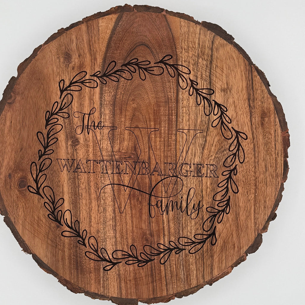 A round wooden plaque featuring the engraved text The Wattenbarger Family inside a decorative wreath design. The letter W is prominent in the background. The wood has a natural grain and a rustic, bark-trimmed edge.
