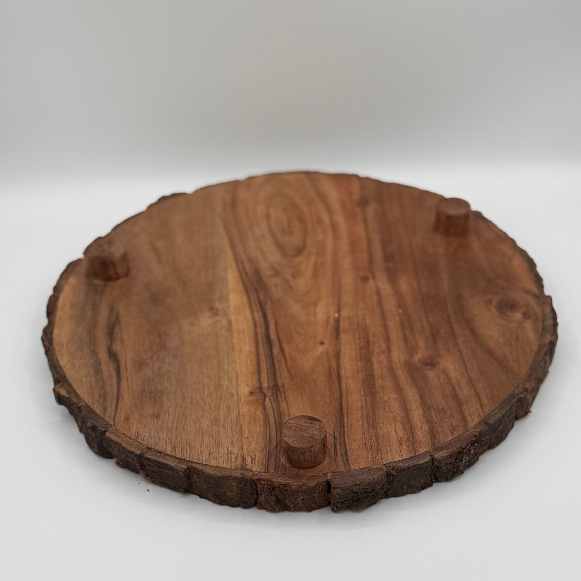 A round wooden serving board with a natural, rustic finish. The board has a smooth surface, visible wood grain, and bark on the edges. Four small wooden feet are attached on the underside, elevating it slightly from the surface. The background is plain white.
