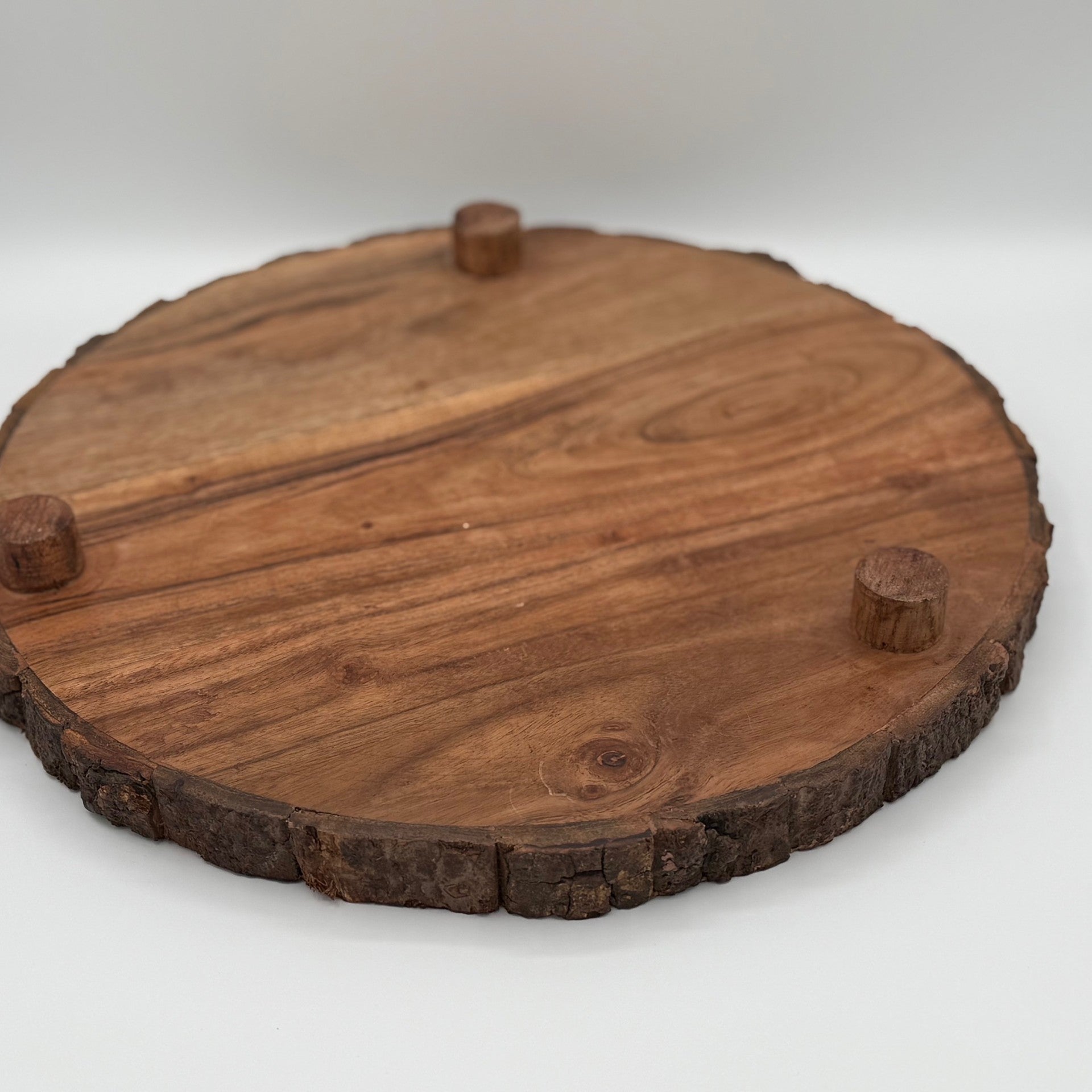 A round wooden board with visible natural grain patterns and tree bark edges sits on a white surface. The board has three small, cylindrical wooden feet attached underneath, elevating it slightly. The wood has a warm, brown tone.