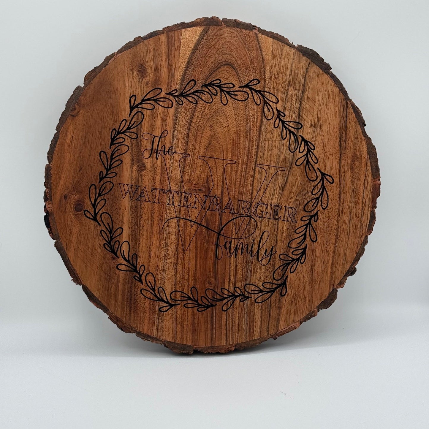 A round wooden sign featuring the engraved text The Whitener Family with a large, decorative letter W behind it. The text is surrounded by an ornate leafy wreath design. The sign has a natural wood finish with visible grain and bark edging.