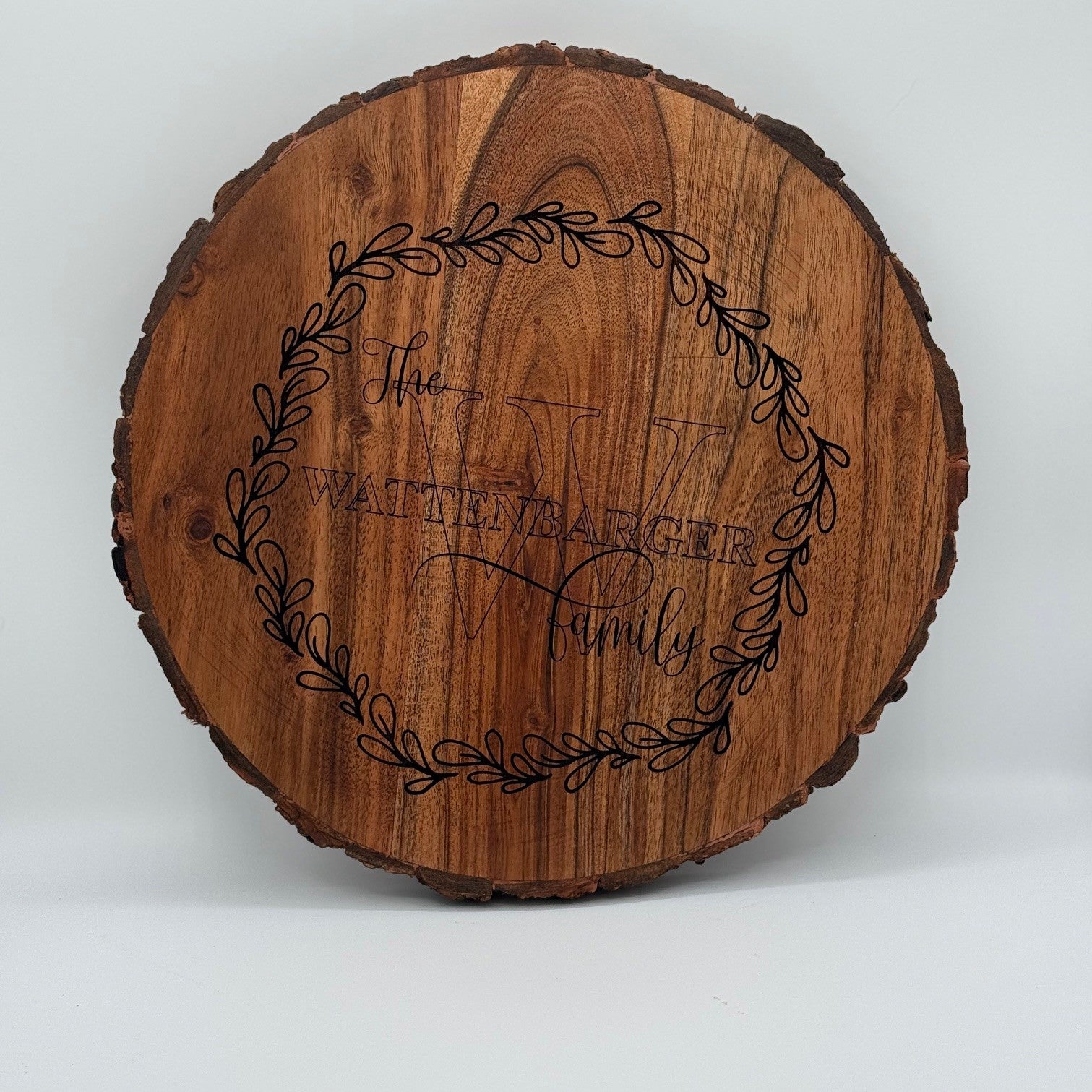 A round wooden sign featuring the engraved text The Whitener Family with a large, decorative letter W behind it. The text is surrounded by an ornate leafy wreath design. The sign has a natural wood finish with visible grain and bark edging.