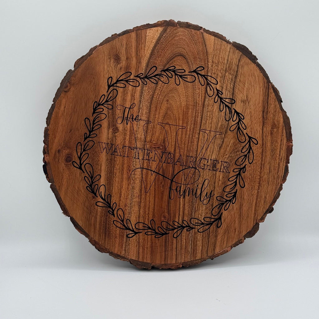 Round wooden plaque with a natural bark edge, featuring the engraved text The Wittenbarger Family over a large letter W. A decorative wreath of leaves encircles the text, creating a rustic, personalized design.
