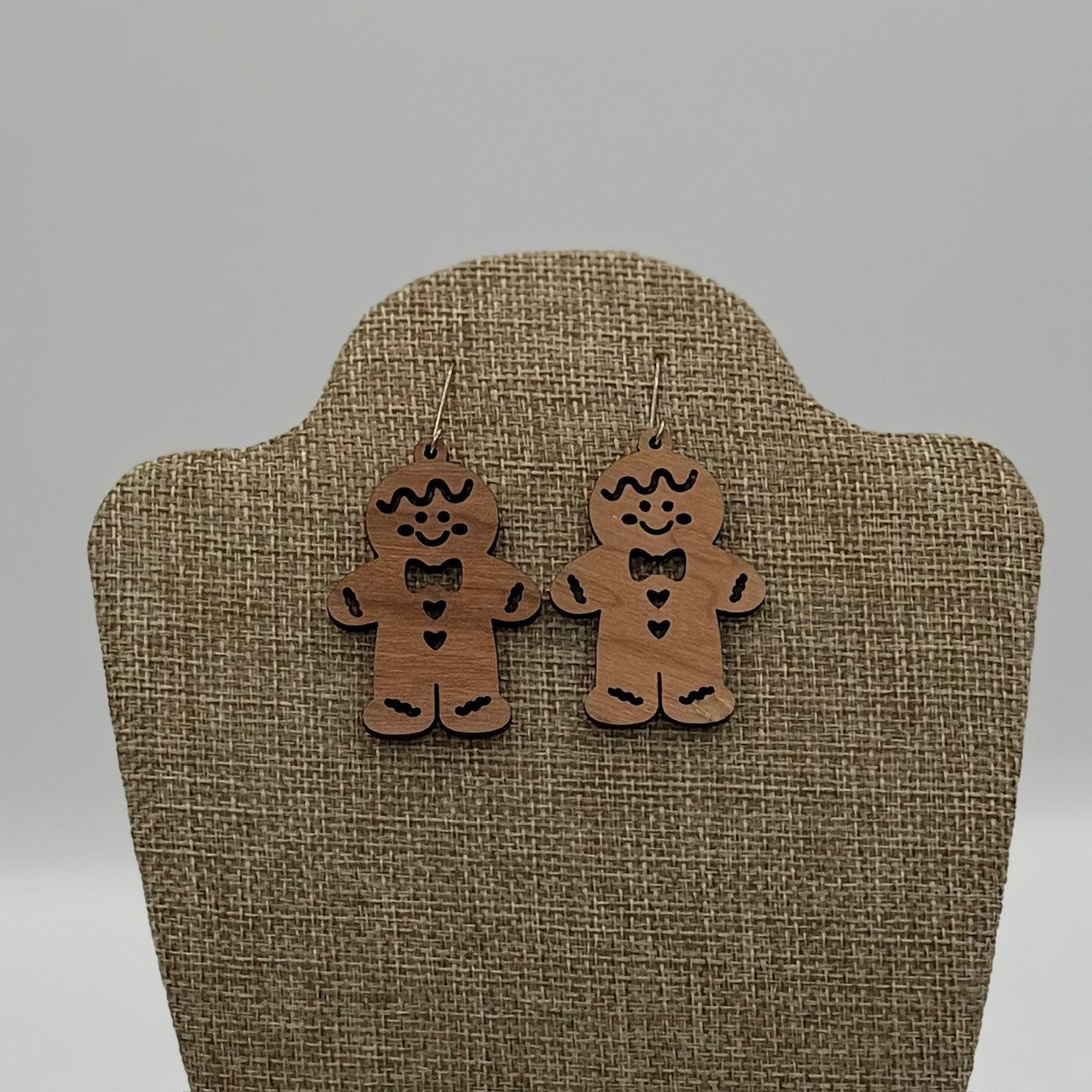 A pair of gingerbread man-shaped wooden earrings displayed on a beige, textured mannequin stand. Each earring features a smiling face, bow tie, and heart detail carved into the wood. The background is light gray.