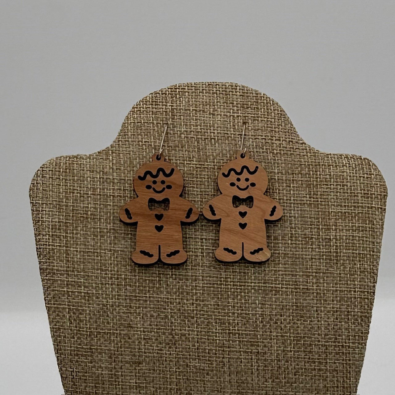 A pair of wooden gingerbread man earrings with painted details, including eyes, mouth, bowtie, buttons, and outline. They hang from hooks on a burlap-textured display stand against a neutral background. Each earring features a heart in the center.