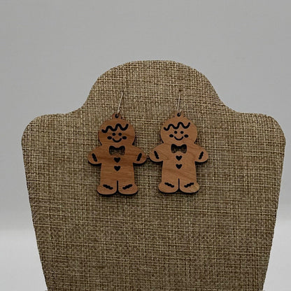 A pair of wooden gingerbread man earrings with painted details, including eyes, mouth, bowtie, buttons, and outline. They hang from hooks on a burlap-textured display stand against a neutral background. Each earring features a heart in the center.
