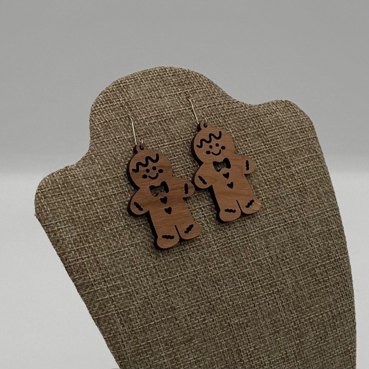 Two wooden earrings shaped like gingerbread men hang on a fabric-covered display stand. The gingerbread men are decorated with bow ties and smiling faces, giving a whimsical and festive appearance. The background is a neutral gray, emphasizing the earthy tones of the earrings.