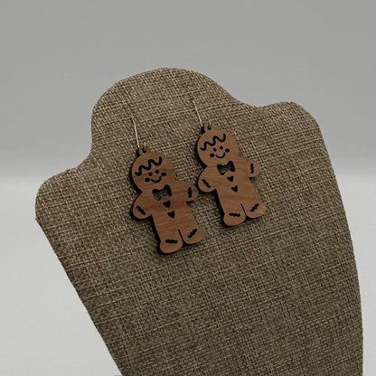 Two wooden earrings shaped like gingerbread men hang on a fabric-covered display stand. The gingerbread men are decorated with bow ties and smiling faces, giving a whimsical and festive appearance. The background is a neutral gray, emphasizing the earthy tones of the earrings.