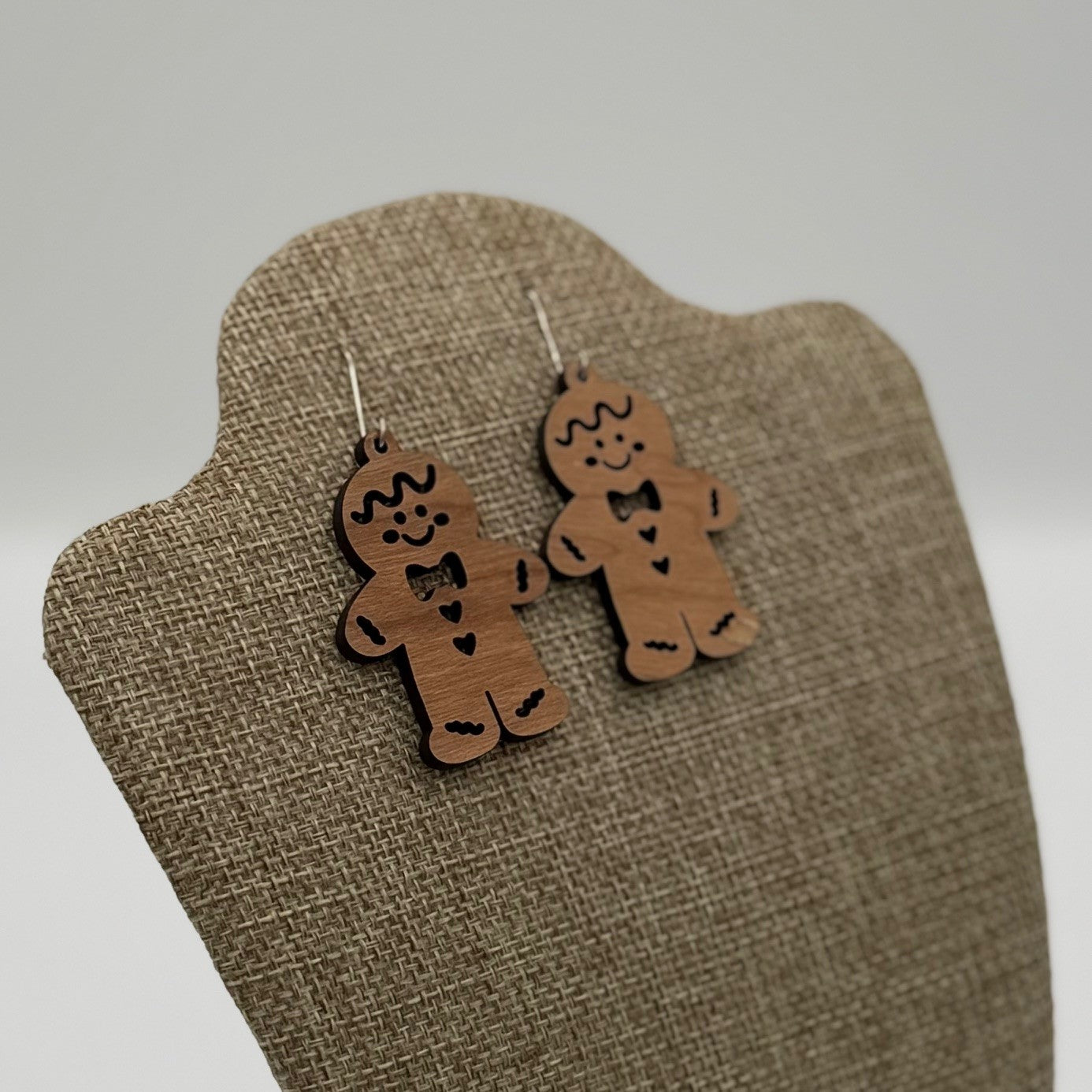 A pair of gingerbread man earrings hangs on a textured beige display stand. The earrings are designed from wood, featuring engraved smiling faces, bow ties, and heart-shaped buttons. The background is softly blurred, highlighting the playful design of the earrings.