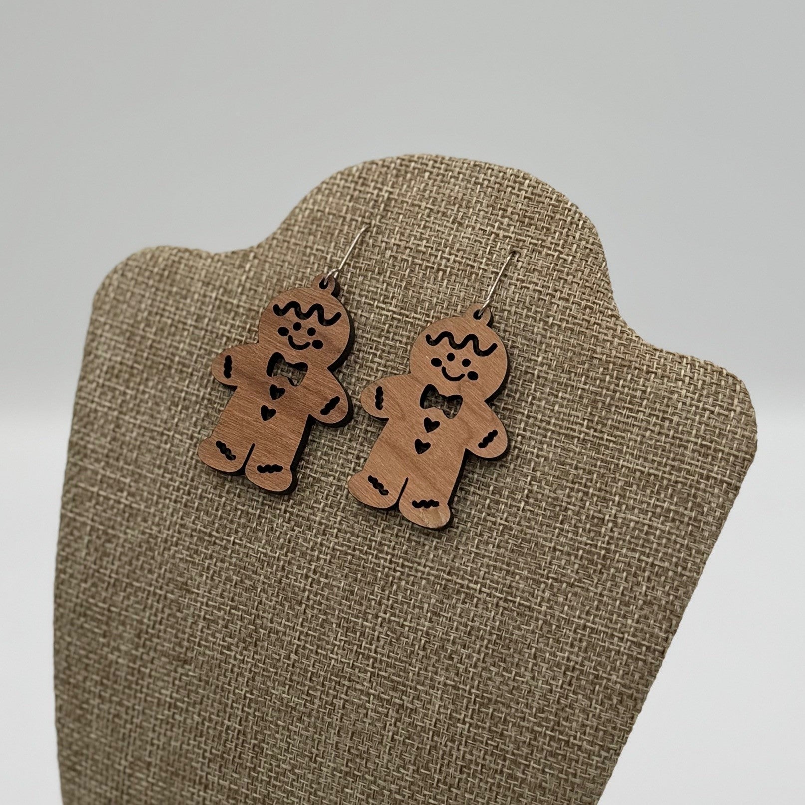 Wooden gingerbread man earrings with heart details hang on a beige textured display stand. The background is soft and blurred, emphasizing the playful, festive design of the earrings.