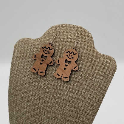 Wooden gingerbread man earrings with heart details hang on a beige textured display stand. The background is soft and blurred, emphasizing the playful, festive design of the earrings.