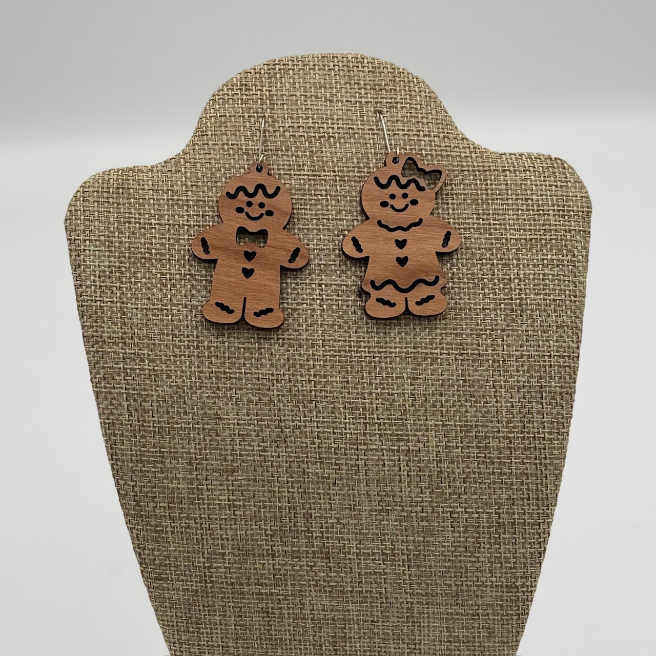 Two wooden gingerbread man earrings are displayed on a textured beige stand. The left earring has a bowtie and buttons, while the right earring features a bow on its head and a heart on the chest. Both are decorated with black accents, giving them a festive, playful appearance.